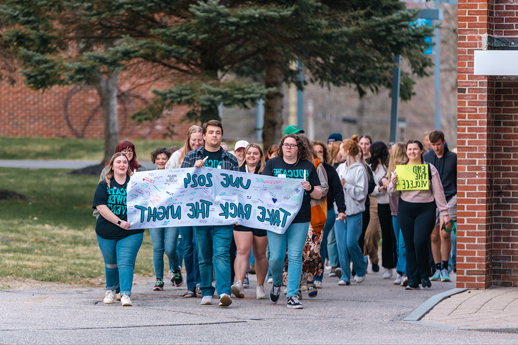 A group of students marches on campus holding a sign that reads "UNE 2024 Take Back the Night"